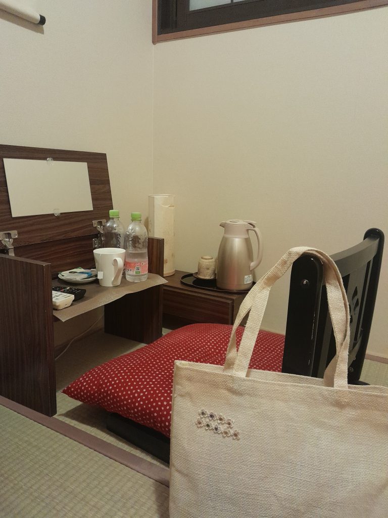 Intérieur d'une chambre chez l'habitant à Kyoto avec un bureau en bois, un miroir et un sac en toile posé sur un fauteuil rouge
